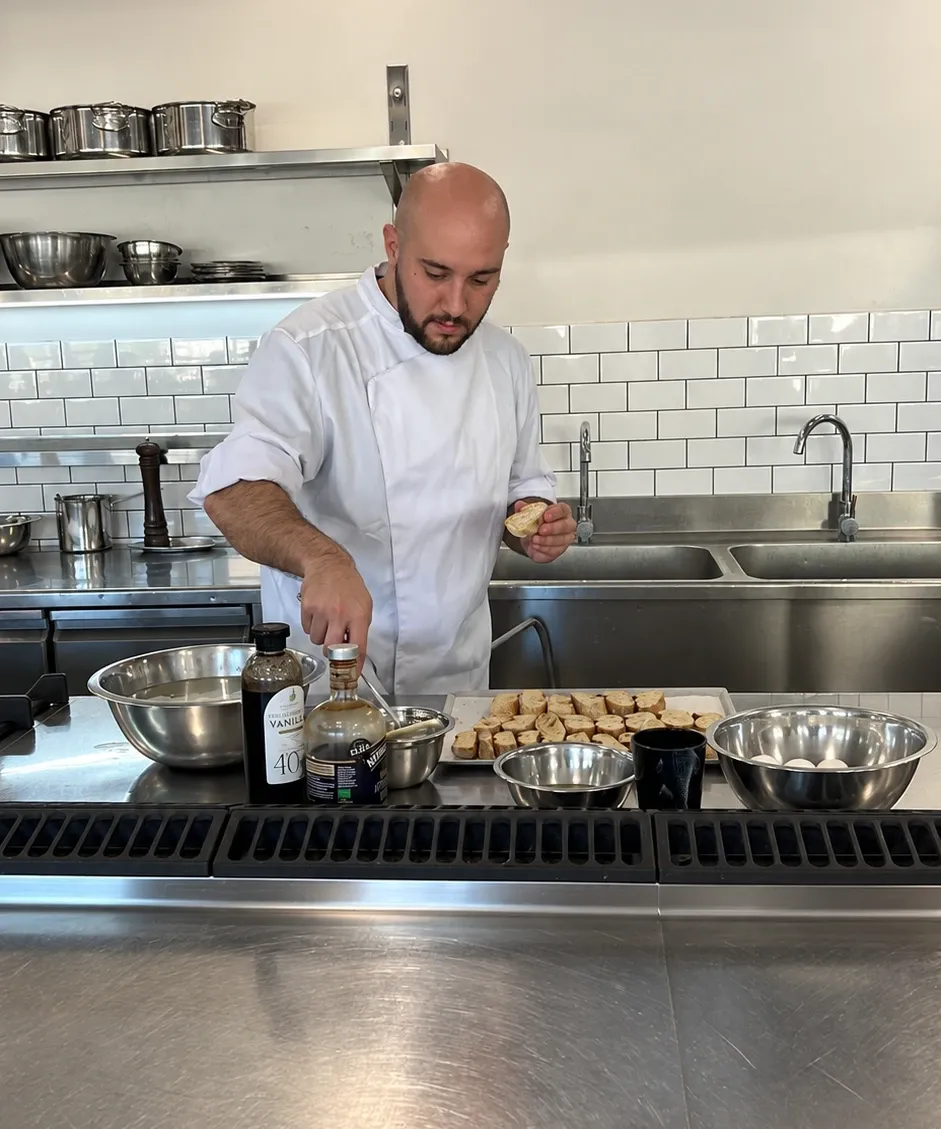 Chef Asaf Zer in a white chef's jacket, holding a slice of bread while he works at a steel counter in a professional kitchen.