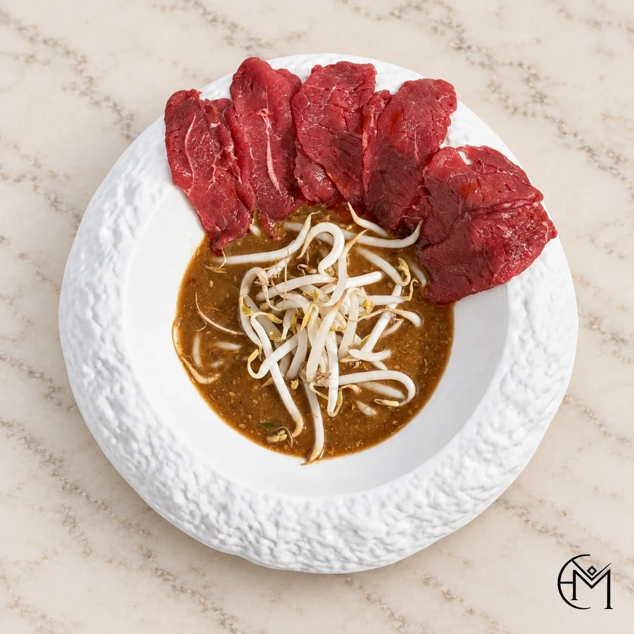 Overhead view of thinly sliced red meat beside a dark broth topped with bean sprouts, in a textured white bowl.