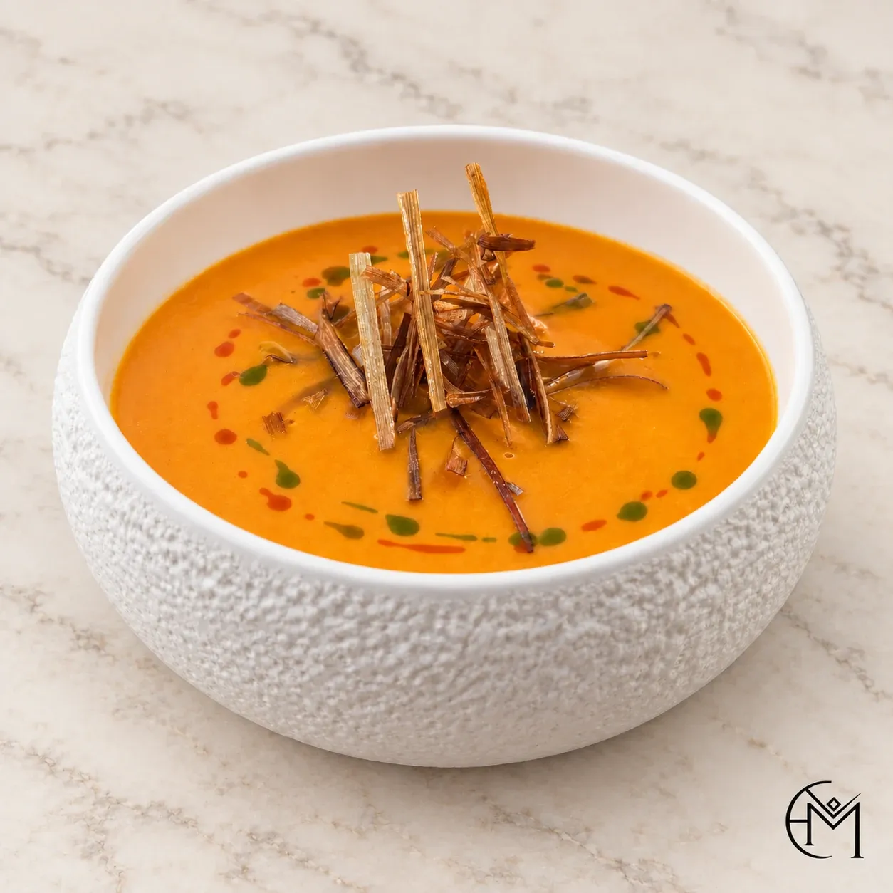 Smooth orange soup topped with a nest of fried strands and dots of red and green oil, in a textured white bowl.