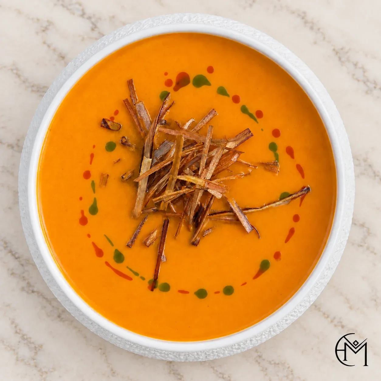 Overhead view of orange soup with a nest of fried strands and a ring of red and green oil.